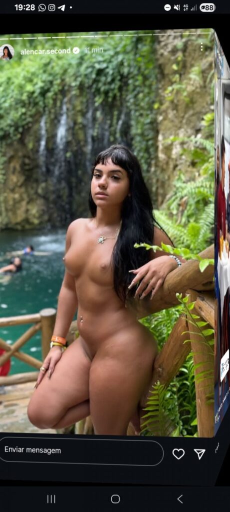 Nude woman posing outdoors by a wooden railing beside a waterfall in a lush setting.